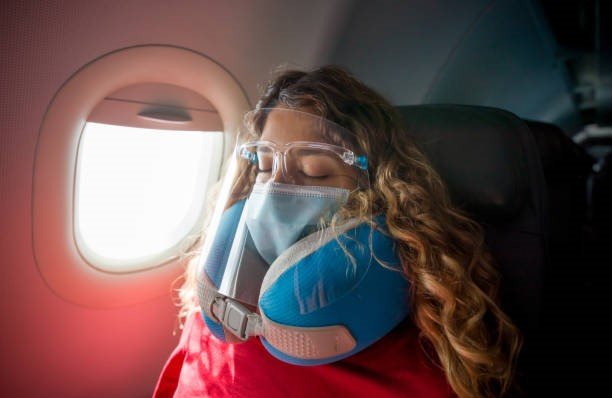 Woman wearing a facemask and a face shield while sleeping in an aeroplane using a neck support pillow 